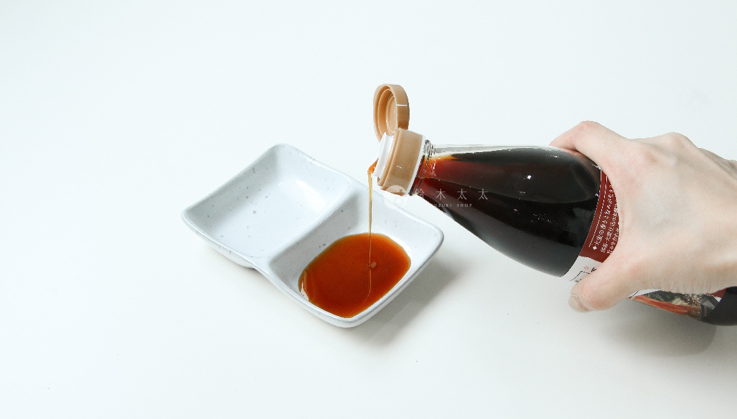 一只手正將一瓶深棕色醬汁倒入一個有兩個隔間的白色陶瓷碟中，碟中已盛有少許醬汁。