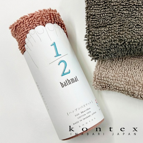 這張照片展示了 KONTEX 品牌的 1/2 迷你快乾吸水浴墊。畫面中心是捲起包裝的粉色浴墊，包裝紙上繪有腳趾圖案以及 1/2 bathmat 字樣，下方標註尺寸為 36cm x 45cm，材質為百分之百純棉，並標明產地為日本今治。背景左上方與右方則分別擺放了深灰色與淺褐色的浴墊，呈現出絨毛般的細緻質感與多種色彩選擇。