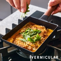 黑色琺瑯鑄鐵玉子燒平底鍋，配備木質手柄，鍋內盛有金黃色湯汁和炸物，上方點綴綠色香草。