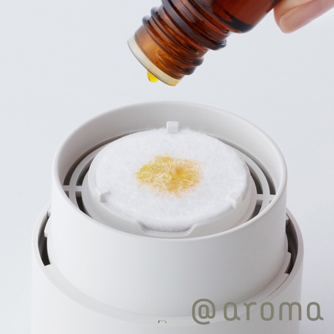 白色圓形替補棉片置於白色香氛機內，一瓶琥珀色玻璃瓶裝精油滴落黃色精油於棉片中央。