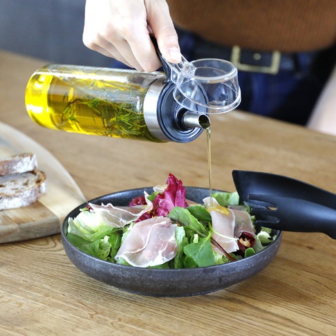 一個女人正在將一個帶有迷迭香的橄欖油瓶傾斜，將油淋在裝滿生菜和義大利火腿的沙拉上。