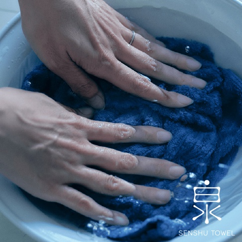 一雙手正在藍色蓬鬆長纖維的浴巾中搓揉，水珠飛濺，浴巾質地細緻，呈現緞澤光澤。