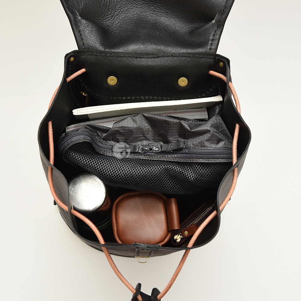黑色皮革單扣束口後背包內部，裝有筆記本、黑色網狀收納袋、金屬水壺、棕色皮革小包和拉鍊錢包。