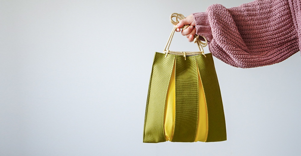 一人穿著淡紫色針織毛衣，手提一只由京都KOSHO設計的ougi帆布信玄袋，包身採用綠色與黃色拼接的褶皺設計，搭配編織繩提把，整體呈現出簡約且具日式風格的質感。