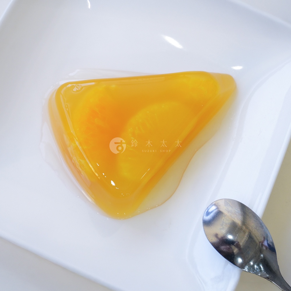 黃色三角形果凍，內含橘色水果切片，果凍表面光滑，盛裝於白色盤中，旁有一銀色湯匙。