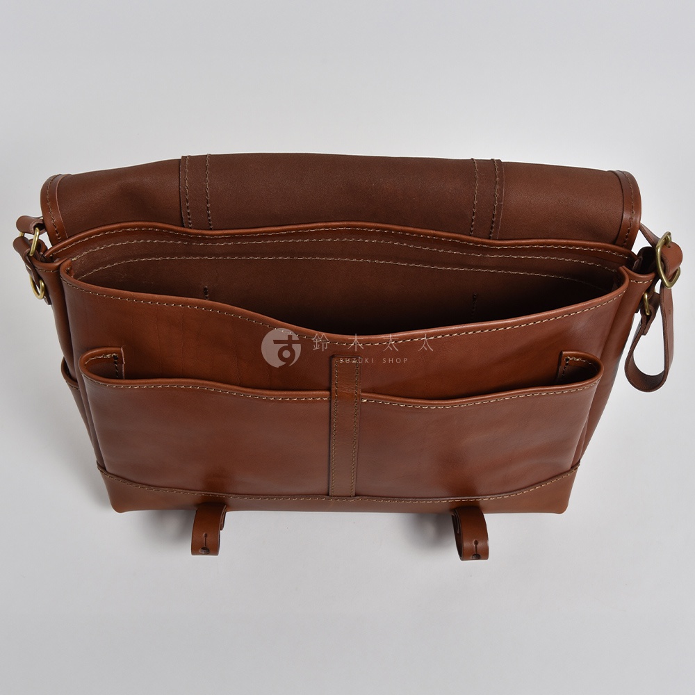職人手作皮革復古郵差包，採用光滑棕色皮革製成，包身有明顯的縫線細節，前方設有兩個開放式口袋，整體呈現經典復古風格。