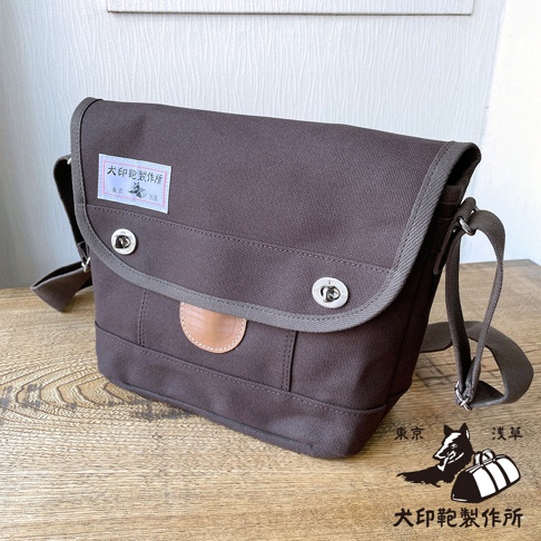 犬印純綿帆布雙扣郵差包，深棕色，配有灰色滾邊和皮革裝飾。包身有兩個銀色旋轉鎖扣，左側有一個印有「犬印製作所」字樣的白色標籤。