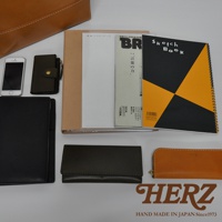 職人手作皮革直式托特包 T-44 L，展示了包含一個手機、一個深綠色皮革零錢包、一個棕色皮革長夾、一個黑色皮革夾、一本筆記本、一本雜誌和一本黑色黃色幾何圖案的 Sketch Book。