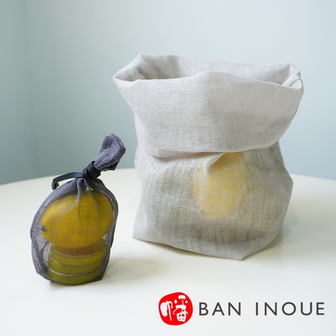 兩個蚊帳布製成的透氣蔬果收納袋，一個為深灰色網眼材質，內裝黃色檸檬，袋口以黑色繩子綁緊；另一個為淺灰色網眼材質，袋口向下翻折，露出袋內黃色蔬果。