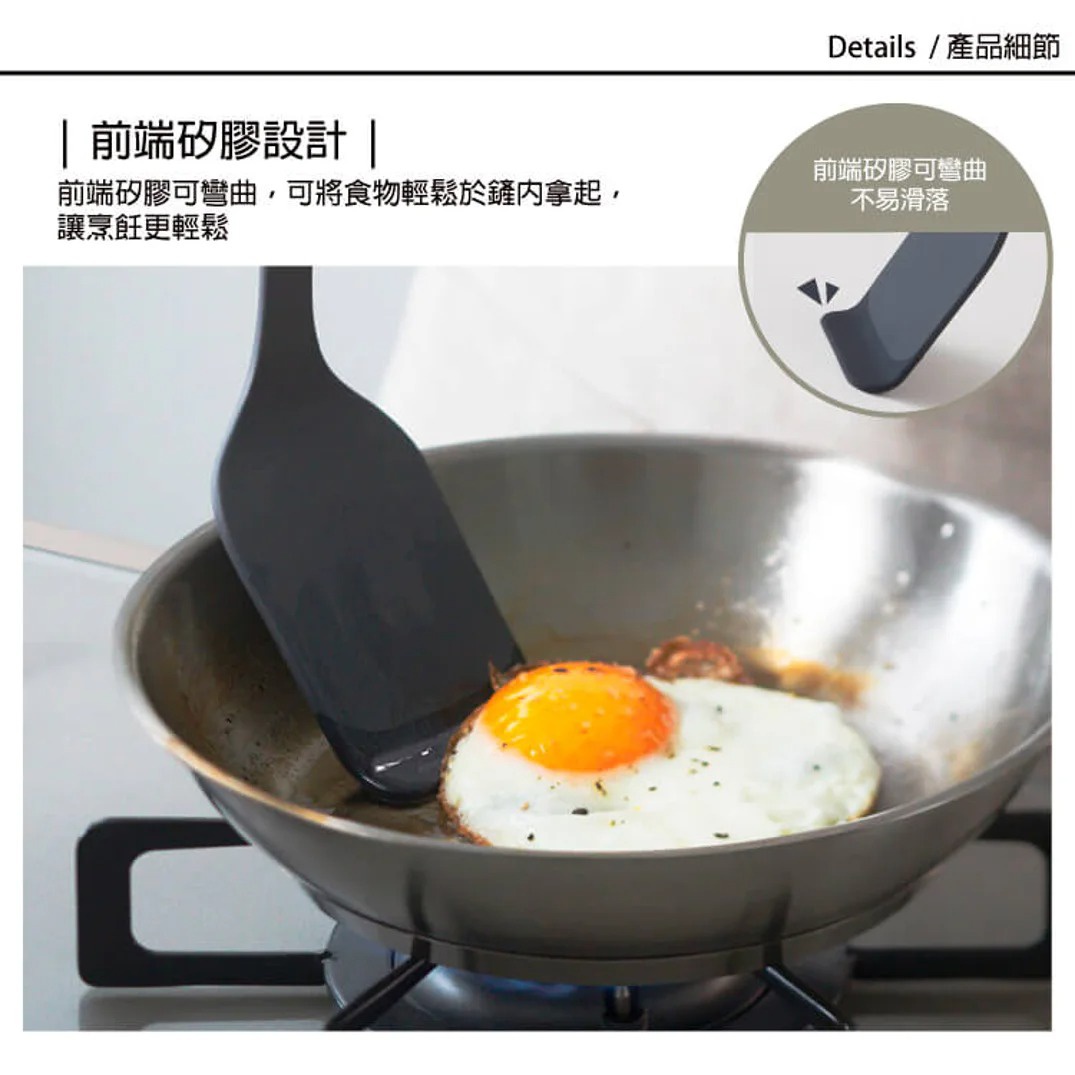 一款黑色的耐熱矽膠鍋鏟正在使用中，它正將一個煎好的荷包蛋從不銹鋼鍋中鏟起，鍋裡還有一些煎蛋的油漬。