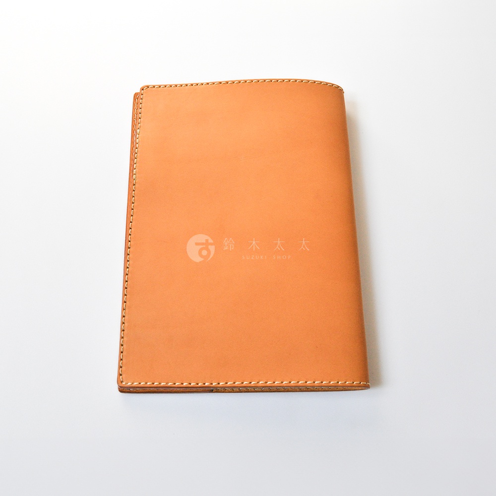 一款淺棕色皮革製成的筆記本書套，採用精緻的縫線工藝，表面帶有鈴木太太的品牌標誌，呈現簡約且富有質感的職人手作風格。