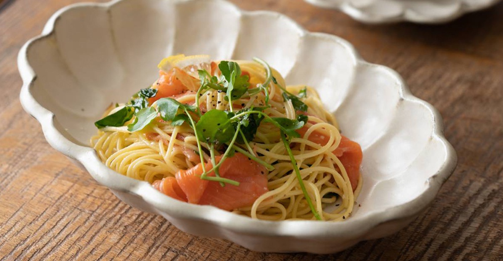 六魯粉引花型橢圓料理盤，白色陶瓷材質，盤緣呈現花瓣狀，盛裝著煙燻鮭魚義大利麵，搭配綠葉裝飾，整體風格簡約溫潤。