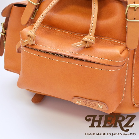 職人手作皮革復古風旅行後背包，採用溫暖的棕褐色皮革，搭配精緻的白色縫線與復古金屬扣環，展現經典質感。背包前方有一個拉鍊小口袋，側邊則有雙口袋設計，整體風格簡約而實用。