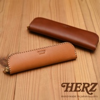 兩個棕色皮革製的筆袋，一個淺棕色，一個深棕色，擺放在木紋桌面上，淺棕色筆袋上有HERZ品牌標誌。