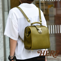 職人手作皮革迷你後背醫生包，橄欖綠色，展示於一名穿著白色襯衫的人背後。包包採用圓潤造型，配有金色金屬扣和提把。