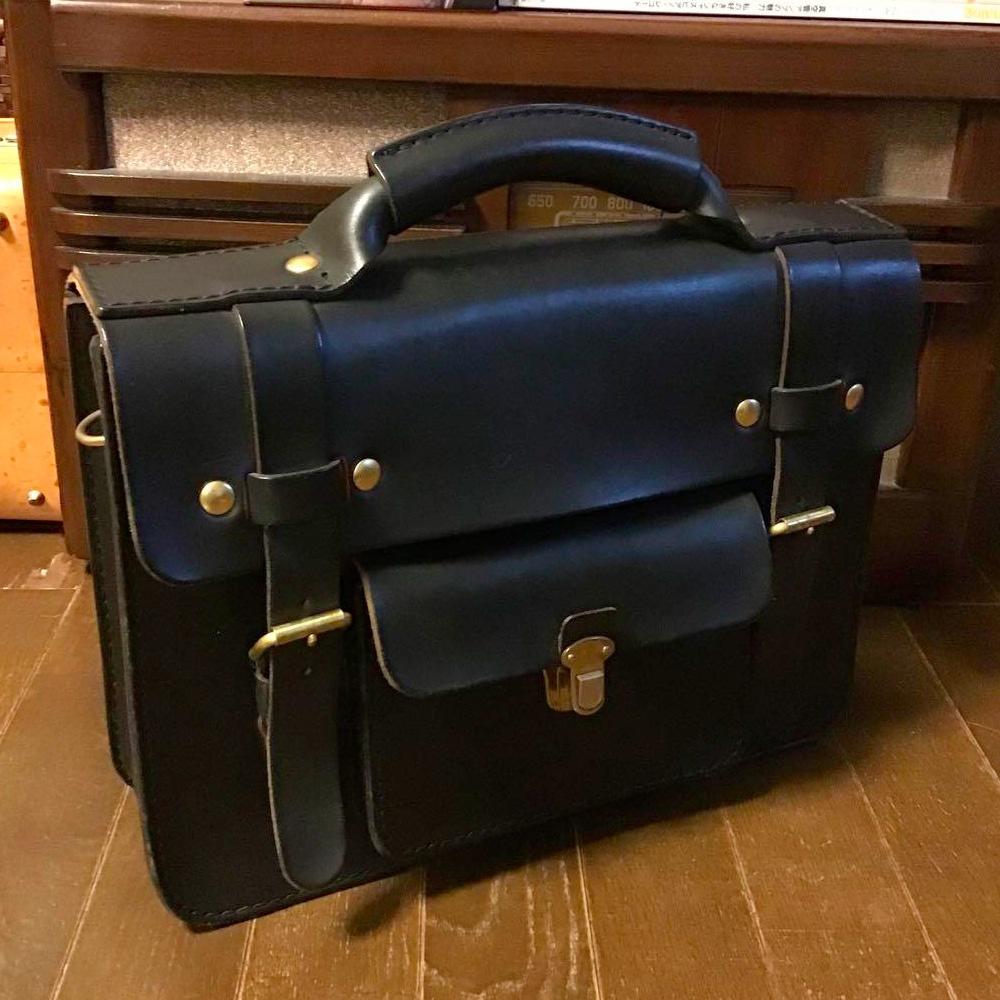 黑色皮革學院風兩用公事包，配有金色金屬扣環和鉚釘，頂部有提把，正面有翻蓋口袋和兩條皮帶固定。