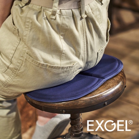 深藍色圓形MINI PUNI Plus坐墊，表面有細微縫線，置於木質圓凳上，一位穿著卡其色褲子的人坐於其上。