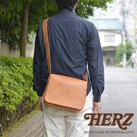 日本HERZ職人手作皮革軟皮橫式斜背包，由側面看，一名男子穿著深藍色襯衫，搭配米白色長褲，斜背著焦糖色皮革斜背包。