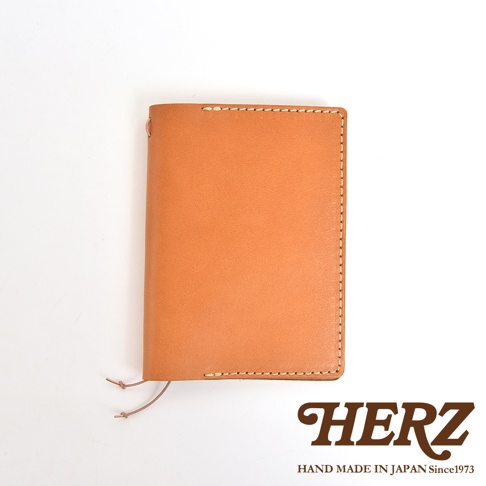 淺棕色皮革手帳套，縫線清晰，右下角有HERZ品牌標誌和HAND MADE IN JAPAN Since1973字樣