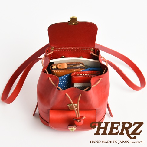 一款日本HERZ手工製作的紅色皮革迷你後背包，採用經典束口設計，包身呈現溫潤的皮革質感。俯視角度展示背包開啟狀態，內部可見放置的錢包、筆記本等小物，包身前方設有翻蓋口袋，底部印有HERZ品牌商標與手工製造資訊。