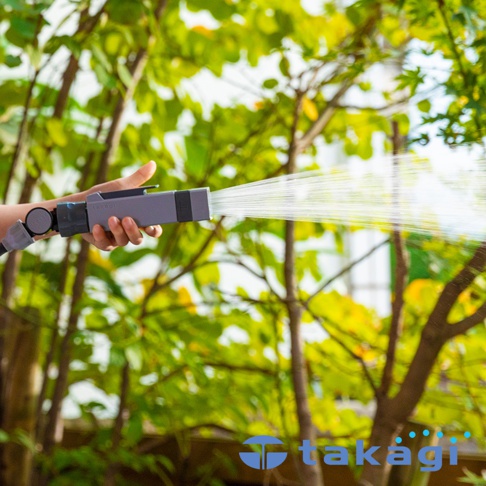 手持灰色與黑色設計的輕量園藝桿型水槍，正在噴射水流，背景為綠意盎然的樹林。