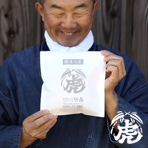 一位男性手持白色包裝的孟宗竹炭產品，包裝上印有灰色「虎」字圖案及日文說明，背景為木紋牆面。