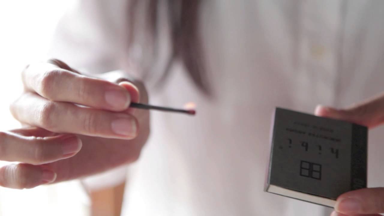 一根點燃的香氛火柴，火光微弱，被一隻手握住。旁邊是一盒深灰色紙質的火柴盒，上面印有黑色文字與圖案。