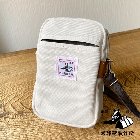 淺米色石洗帆布材質的斜背手機包，正面中央有紅色邊框的白色方形布標，印有黑色圖案及日文文字，包身側邊有棕色皮革飾條，搭配深棕色斜背帶。