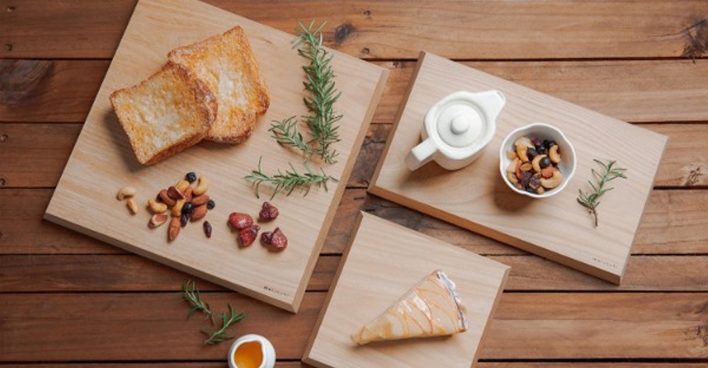 榆木砧板上擺放著吐司、堅果、草莓乾、茶壺和一塊蛋糕，周圍點綴著迷迭香。