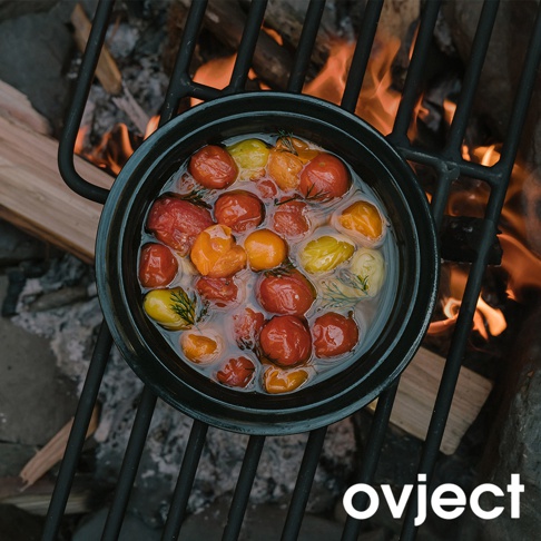 黑色琺瑯湯鍋置於烤架上，鍋內裝有煮熟的紅色、橘色和黃色小番茄，以及香草。鍋下方有燃燒的火焰和木柴。