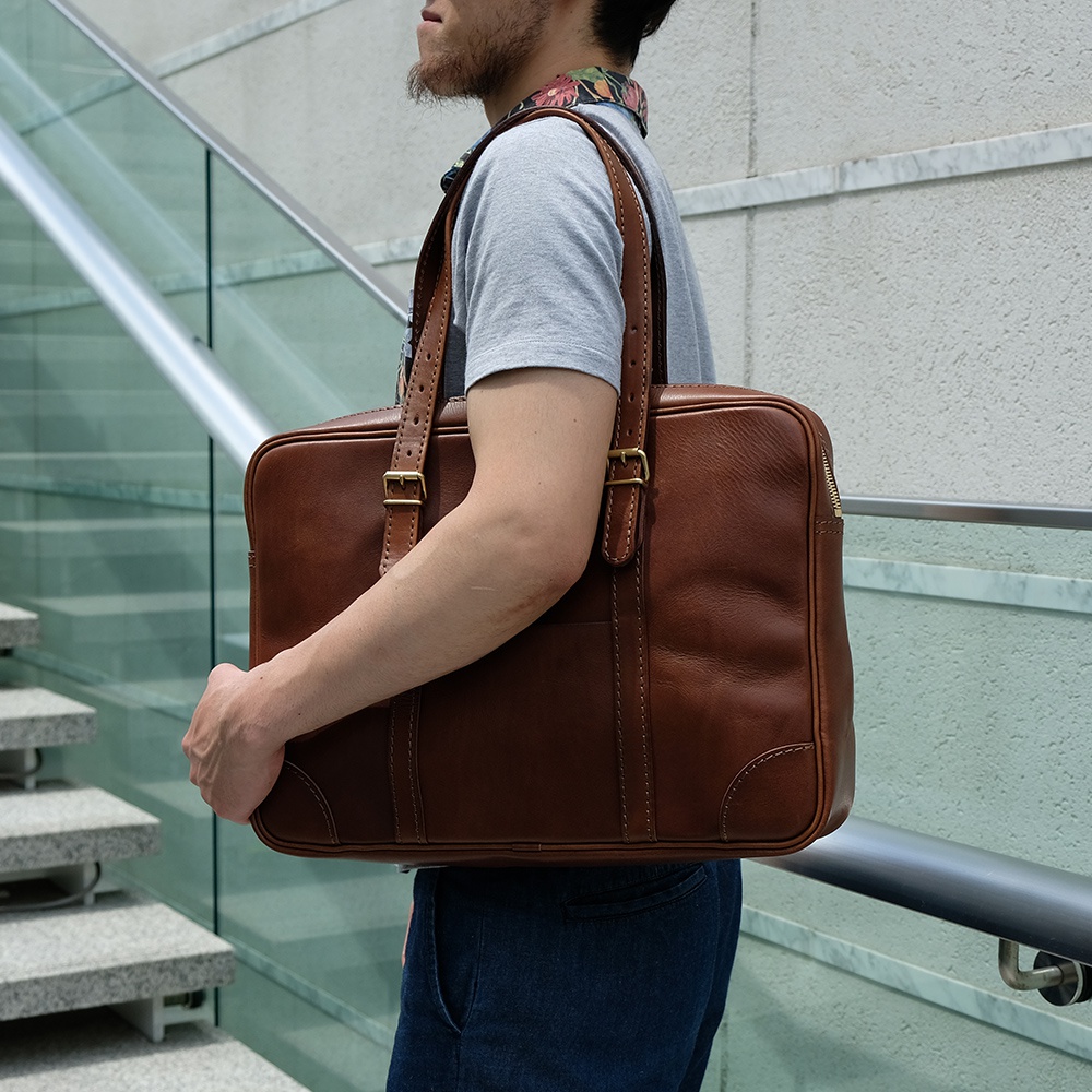 職人手作皮革肩背手提兩用公事包，呈現經典棕色，以光滑牛皮製成，搭配金屬拉鍊與扣環。包款線條俐落，縫線精緻，設計簡約大方。
