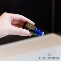 手持一瓶樟楠木條精油，藍色玻璃瓶身，金色標籤印有「KISU HANDM」字樣，瓶蓋處有白色滴管。