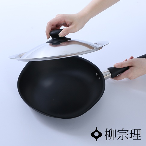 一雙手正拿著不鏽鋼鍋蓋與黑色柳宗理岩紋感單手鐵鍋，鐵鍋呈現獨特的深色質感，鍋身連接黑色長柄，並標註有柳宗理品牌名稱，整體展現簡約實用的廚具設計風格。