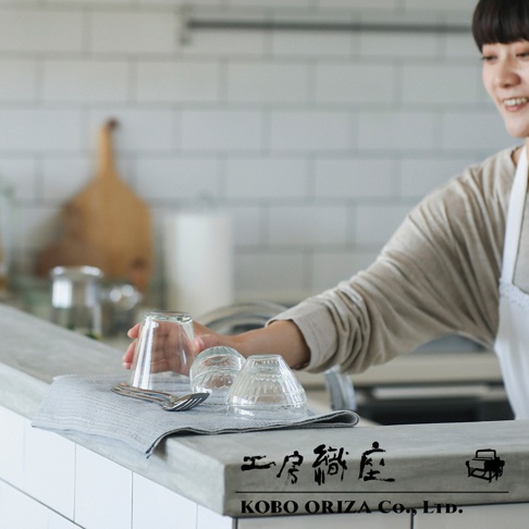 一名女子正在廚房用抹布擦拭玻璃杯，地上有四個玻璃杯和兩支餐具，牆上掛著一個木製掛板。