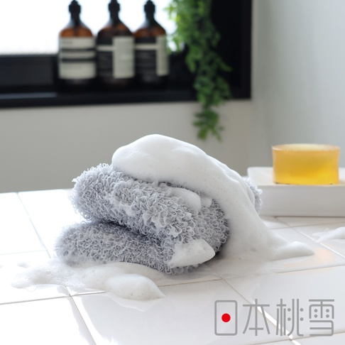 灰色沐浴巾堆疊，覆蓋豐富泡沫，產品置於白色磁磚上，背景可見三瓶深色沐浴乳與一塊黃色肥皂。