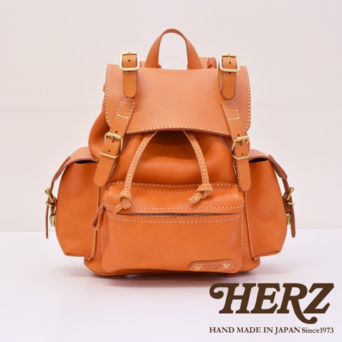 職人手作皮革復古風旅行後背包，採用光滑的棕褐色皮革製成，配有金色搭扣和抽繩閉合。背包前方有拉鍊口袋和品牌標誌。