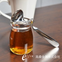 日製不鏽鋼可掛式蜂蜜醬料匙，銀色，勺柄呈彎曲狀，可掛置於玻璃罐邊緣，勺頭裝有琥珀色液體。