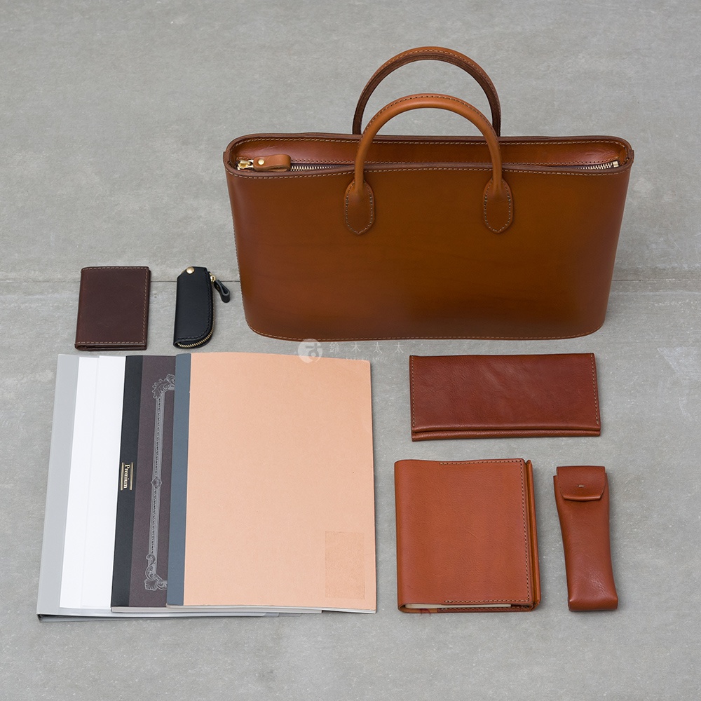 職人手作皮革Organ薄型公事包，以棕色皮革製成，配有雙提把和拉鍊開口。周圍展示多個皮革配件，包括卡夾、鑰匙包、筆記本、長夾和筆套。