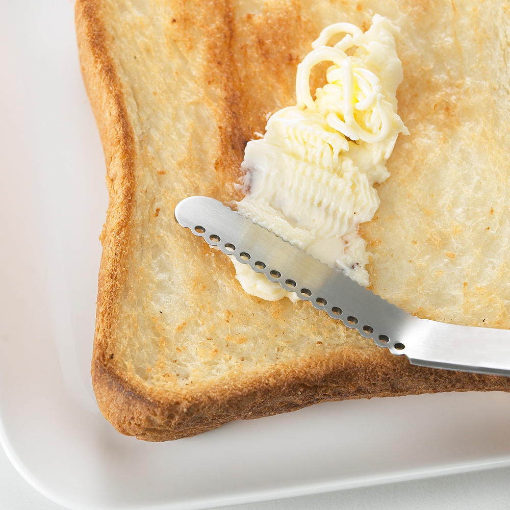 日製奶油刀，不鏽鋼材質，刀刃帶有小圓孔鋸齒，正塗抹奶油於烤吐司上。