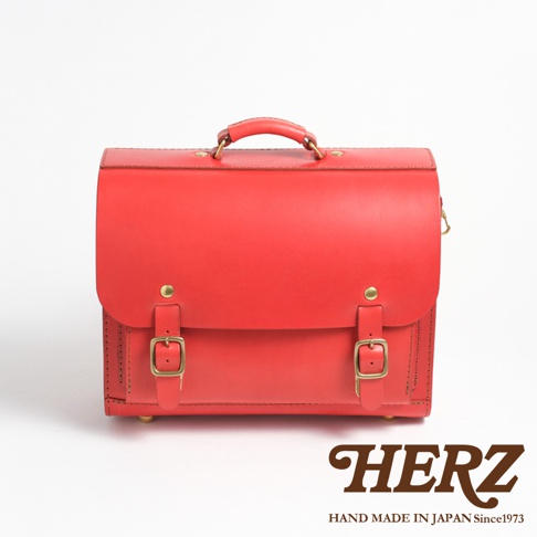 一個鮮紅色的皮革橫式書包，具有頂部提把、兩個前方的皮帶扣和金屬腳釘。書包的右下方印有HERZ字樣和HAND MADE IN JAPAN Since1973的字樣。