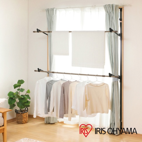 木紋頂天立地窗框曬衣架，黑色立柱，兩層掛桿，上方掛有兩條白色毛巾，下方掛有七件不同顏色（白色、灰色、米色）的長袖衣物，安裝於窗戶旁。