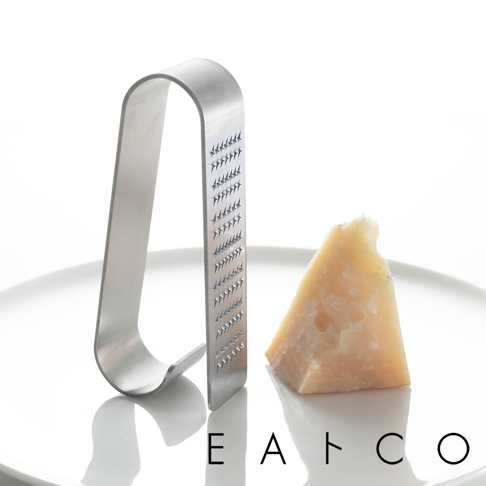 日製立式磨泥器，採用銀色金屬材質，呈U字形設計，一側有密集的磨齒。旁邊擺放一塊三角形的起司，一同置於白色盤上。