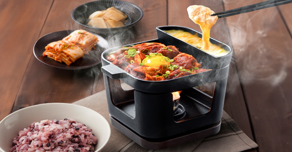 黑色鑄鐵烤盤鍋爐組，內有燉煮的肉塊、泡菜和一顆生蛋黃，旁有融化的起司，下方有火焰加熱，整體散發出蒸氣。