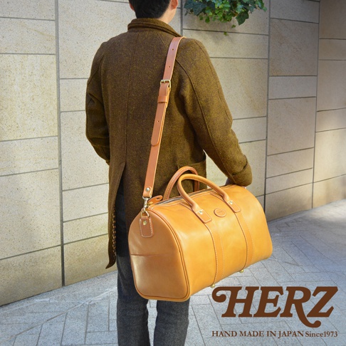 淺棕色皮革製成的職人手作波士頓包，可肩背或手提，配有可調節長度的皮革背帶和兩個圓形提把，包身拉鍊開合，側邊有皮革飾條和縫線細節。