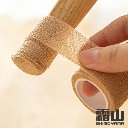 一隻手正將米色、網狀紋理的纏繞式膠帶纏繞在木製桌椅腳上，膠帶具有彈性，能緊密貼合，防止刮傷並減少噪音。