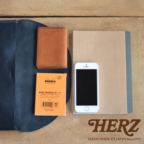 職人手作皮革小物收納袋，展示深藍色皮革外層，內有咖啡色皮革卡夾、白色手機及黃色筆記本，置於木質背景上。