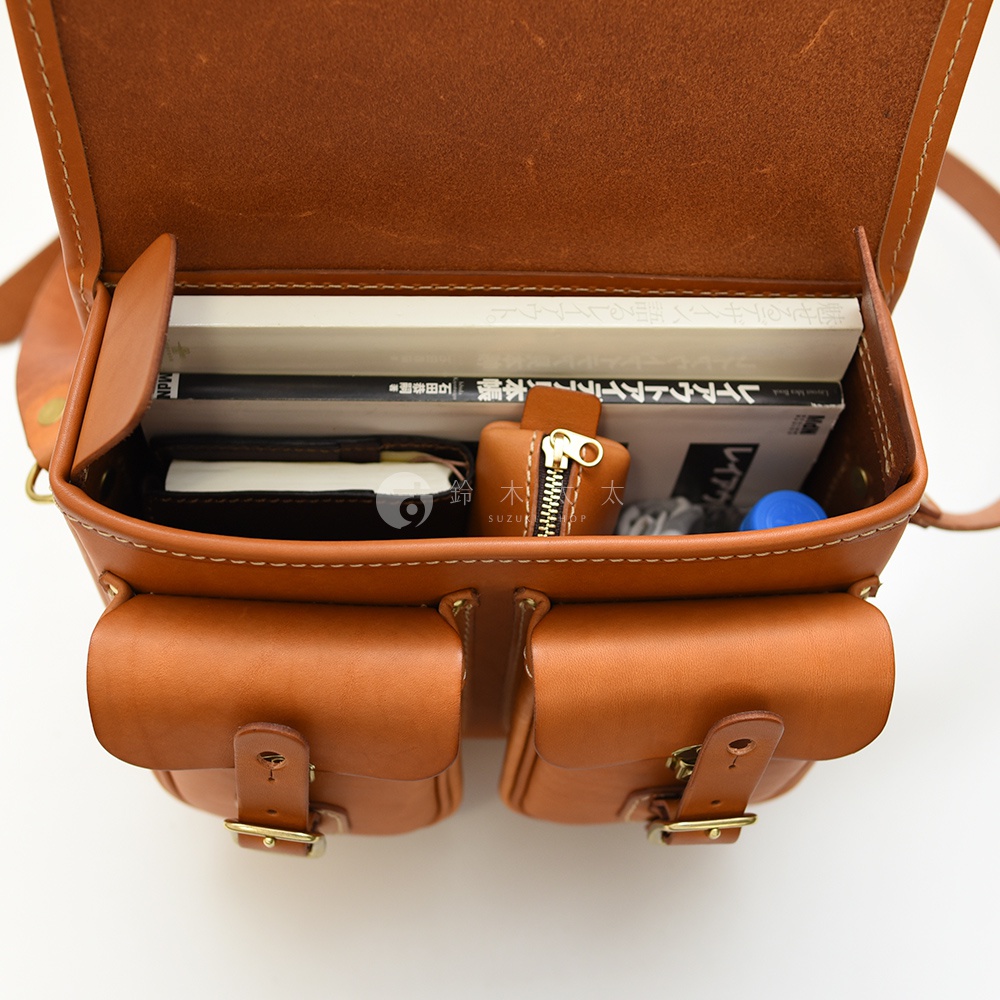 HERZ 職人手作皮革雙扣郵差包內部的商品，包含書籍、筆記本和一個鑰匙包，頂部打開展示了包包的內部空間。