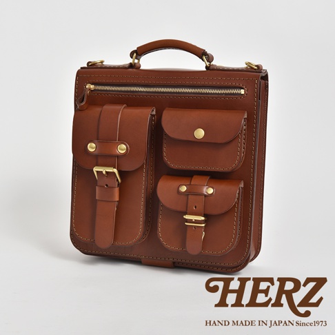 職人手作棕色皮革三用後背包，設有三個前袋，其中兩個帶有金屬扣帶和搭扣，一個帶有金屬鈕扣。背包頂部有拉鍊開合和皮革提把，整體風格簡潔實用。