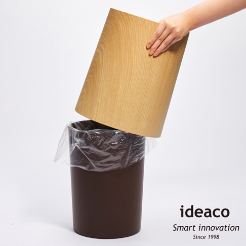 一個深棕色圓柱形垃圾桶，內有透明垃圾袋，上方有一個橡木紋的圓筒形蓋子，正被一隻手提起。