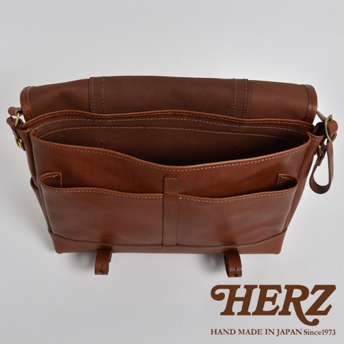 職人手作皮革復古郵差包，採用經典棕色，以精緻縫線勾勒出包身線條，正面設有兩個貼袋，整體呈現質感與實用兼具的設計。